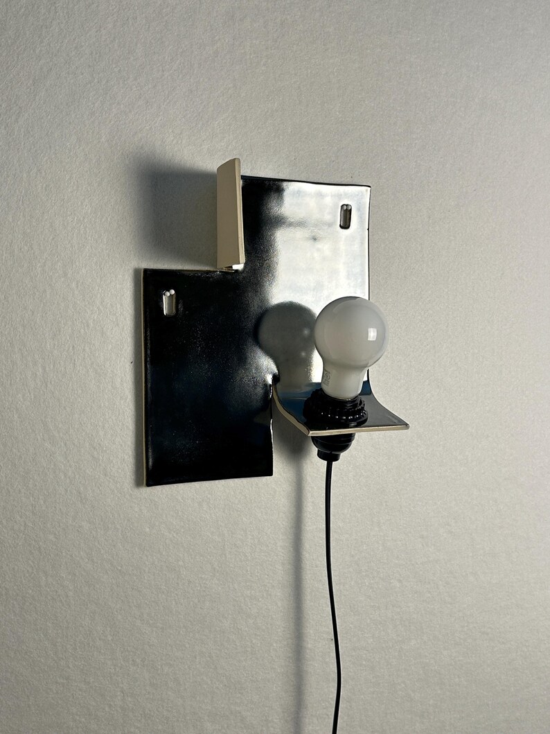 Pode incluir: Uma lumin&aacute;ria de parede moderna com uma tela geom&eacute;trica de metal preto e uma l&acirc;mpada branca. A lumin&aacute;ria tem um design minimalista com uma forma retangular e um pequeno detalhe em branco sujo. Um cabo preto se estende da l&acirc;mpada.