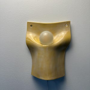 May include: A wall-mounted ceramic light fixture with a glossy, pale yellow finish. The piece features a curved, organic shape with a central indentation holding a single, round, frosted white light bulb. Two small holes are visible at the top.