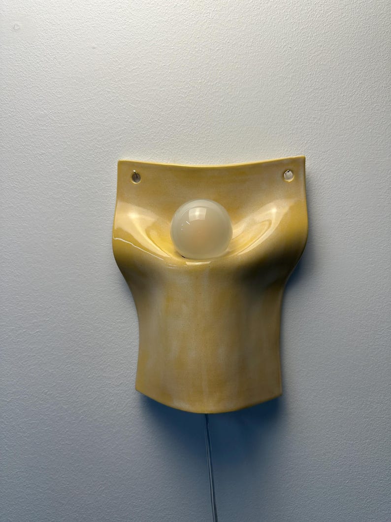 May include: A wall-mounted ceramic light fixture with a glossy, pale yellow finish. The piece features a curved, organic shape with a central indentation holding a single, round, frosted white light bulb. Two small holes are visible at the top.