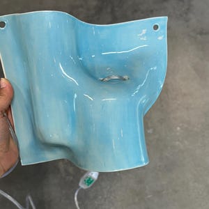 May include: A light blue ceramic wall sconce with a wavy, organic shape and a glossy finish. The fixture features two circular mounting holes at the top corners and a central opening containing a silver metal ring. A white electrical cord is attached.