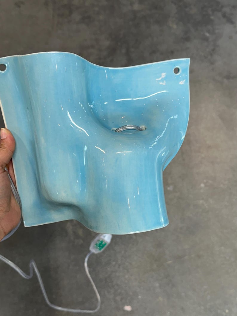 May include: A light blue ceramic wall sconce with a wavy, organic shape and a glossy finish. The fixture features two circular mounting holes at the top corners and a central opening containing a silver metal ring. A white electrical cord is attached.