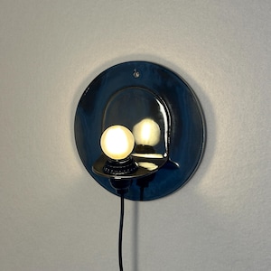 May include: A circular, dark blue glazed ceramic wall sconce featuring a small, protruding shelf that holds a single, glowing spherical light bulb. A thin black electrical cord extends downward from the base of the light fixture.