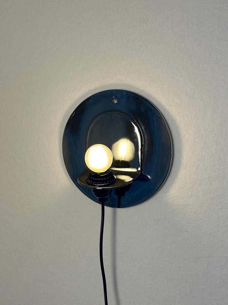 May include: A circular, dark blue glazed ceramic wall sconce featuring a small, protruding shelf that holds a single, glowing spherical light bulb. A thin black electrical cord extends downward from the base of the light fixture.