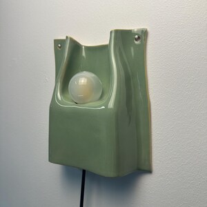 May include: A sage green ceramic wall sconce with a glossy finish and a curved, folded silhouette. The fixture features a central circular opening holding a round, frosted light bulb and includes two small holes at the top for mounting.
