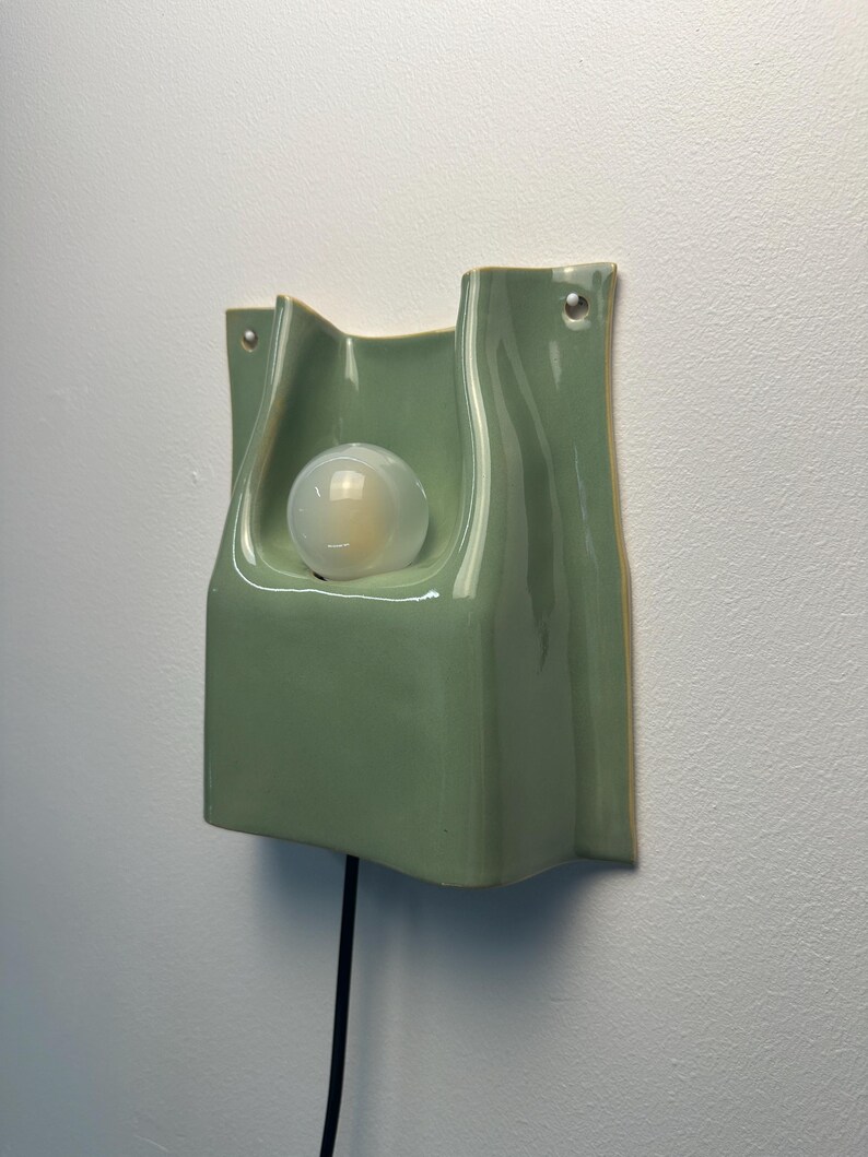 May include: A sage green ceramic wall sconce with a glossy finish and a curved, folded silhouette. The fixture features a central circular opening holding a round, frosted light bulb and includes two small holes at the top for mounting.