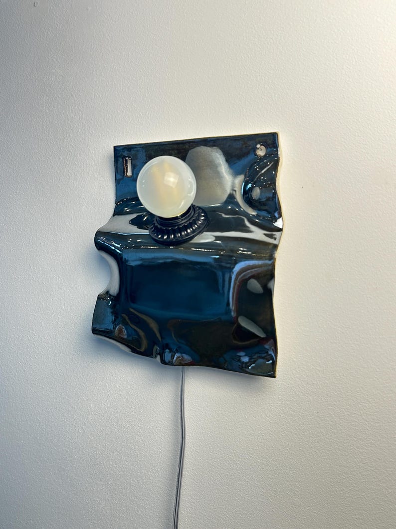 May include: A wall-mounted lamp featuring a glossy, dark, wavy ceramic backplate with a reflective surface. A single round, frosted white light bulb is centered on the front, attached to a black circular base. A thin power cord hangs downward.