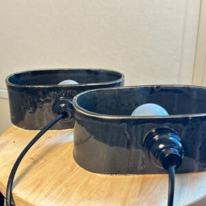May include: Two black ceramic lamps with an oval shape, each with a light bulb inside. The lamps have a glossy finish and are connected to a black cord. They are placed on a wooden stool.