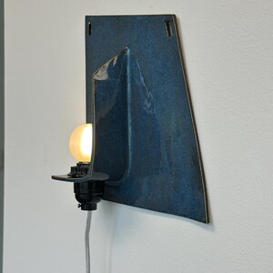 May include: A modern wall sconce with a dark blue ceramic shade. The shade has a unique, folded design and a glossy finish. A black light fixture holds a round, glowing light bulb. A gray power cord hangs down.