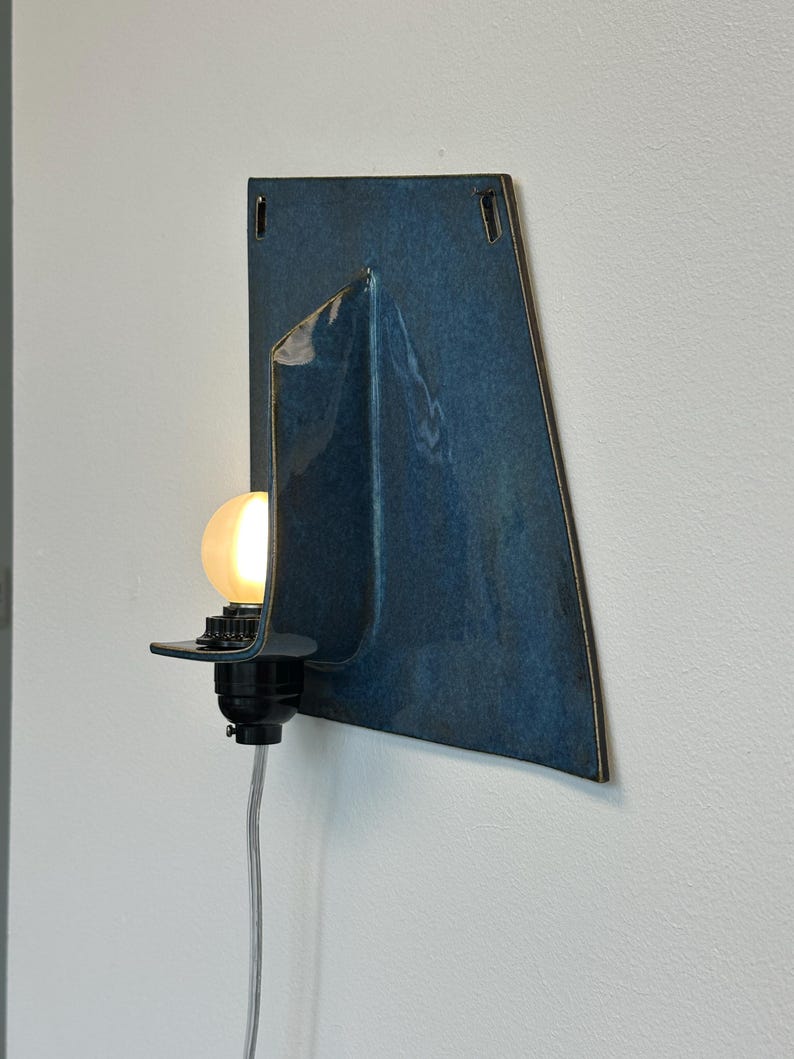 May include: A modern wall sconce with a dark blue ceramic shade. The shade has a unique, folded design and a glossy finish. A black light fixture holds a round, glowing light bulb. A gray power cord hangs down.
