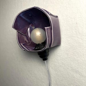 K&ouml;nnte beinhalten: Eine Wandleuchte mit einem gl&auml;nzenden, tiefvioletten Keramikschirm mit unregelm&auml;&szlig;ig gefalteten Kanten. Eine runde, mattwei&szlig;e Gl&uuml;hbirne ist mittig in einer schwarzen Fassung platziert, die mit einem transparenten Stromkabel verbunden ist.