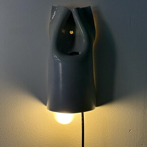 May include: A wall-mounted light fixture with a smooth, glossy, light gray ceramic shade featuring a folded, organic shape. The fixture emits a warm, downward-facing glow from a single bulb, with a black power cord extending from the bottom.