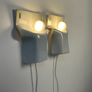 May include: A pair of wall-mounted light fixtures featuring a smooth, glossy white ceramic body with a curved, sculptural form. Each fixture holds a single glowing spherical light bulb, with a thin white power cord extending from the bottom.