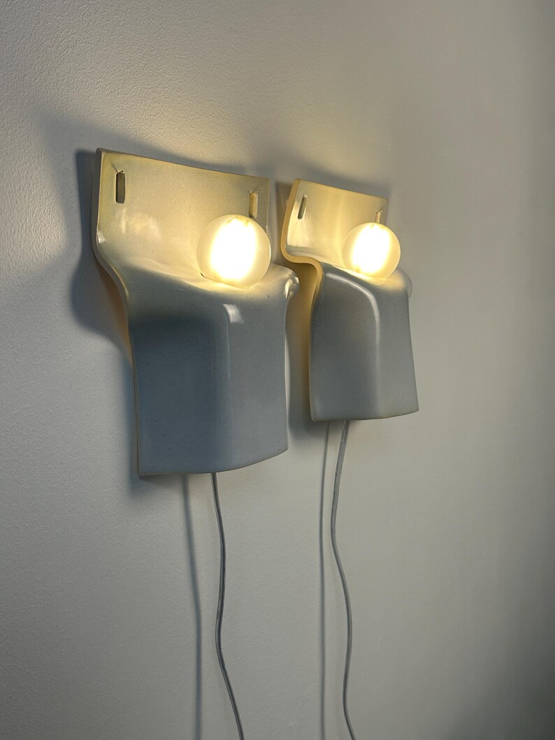May include: A pair of wall-mounted light fixtures featuring a smooth, glossy white ceramic body with a curved, sculptural form. Each fixture holds a single glowing spherical light bulb, with a thin white power cord extending from the bottom.