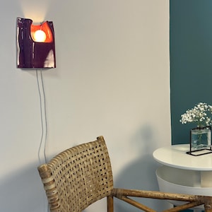 Puede incluir: Un aplique de pared de cer&aacute;mica con un exterior de color morado oscuro brillante y un interior de color naranja vivo. La l&aacute;mpara presenta una forma org&aacute;nica curva con una bombilla redonda que emite una luz c&aacute;lida, conectada por un cable blanco.