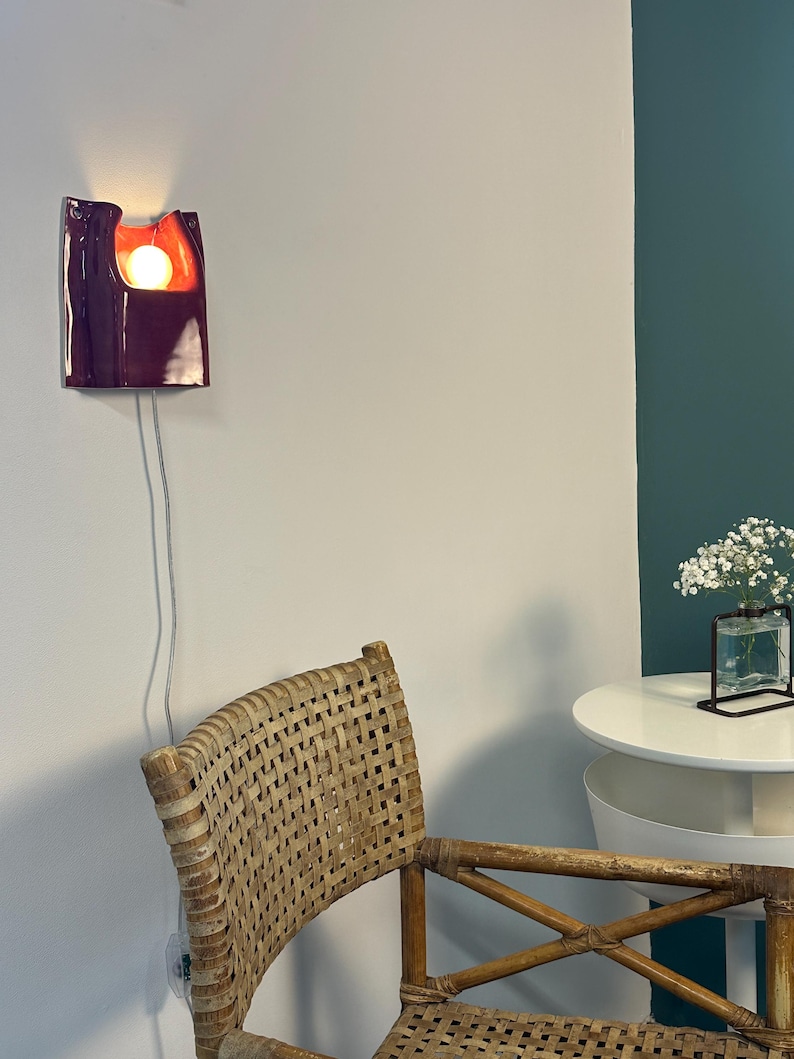 Puede incluir: Un aplique de pared de cer&aacute;mica con un exterior de color morado oscuro brillante y un interior de color naranja vivo. La l&aacute;mpara presenta una forma org&aacute;nica curva con una bombilla redonda que emite una luz c&aacute;lida, conectada por un cable blanco.