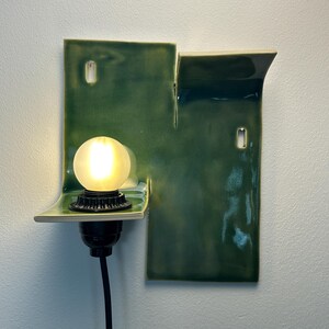 May include: A wall-mounted ceramic light fixture with a glossy, deep green glaze. The structure features two rectangular panels of varying heights with curved edges. A black socket holds a glowing, frosted spherical light bulb, with a black cord.