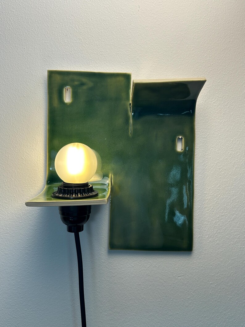 May include: A wall-mounted ceramic light fixture with a glossy, deep green glaze. The structure features two rectangular panels of varying heights with curved edges. A black socket holds a glowing, frosted spherical light bulb, with a black cord.