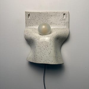 Może przedstawiać: Ścienna oprawa oświetleniowa z ceramiki o białej podstawie w czarne kropki. Lampa ma zakrzywiony, organiczny kształt z centralnym okrągłym otworem, w kt&oacute;rym znajduje się matowa żar&oacute;wka. Czarny przew&oacute;d zasilający zwisa z dolnej części oprawy.