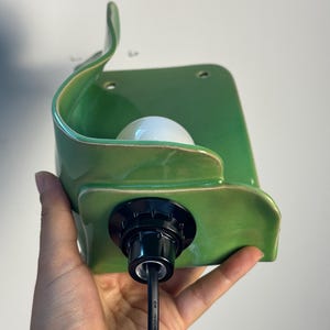 Pu&ograve; includere: Una lampada da parete in ceramica verde con una silhouette curva a forma di foglia e una finitura lucida. L'apparecchio presenta un portalampada centrale bianco e un cavo elettrico nero che esce dalla base, con due piccoli fori di montaggio.