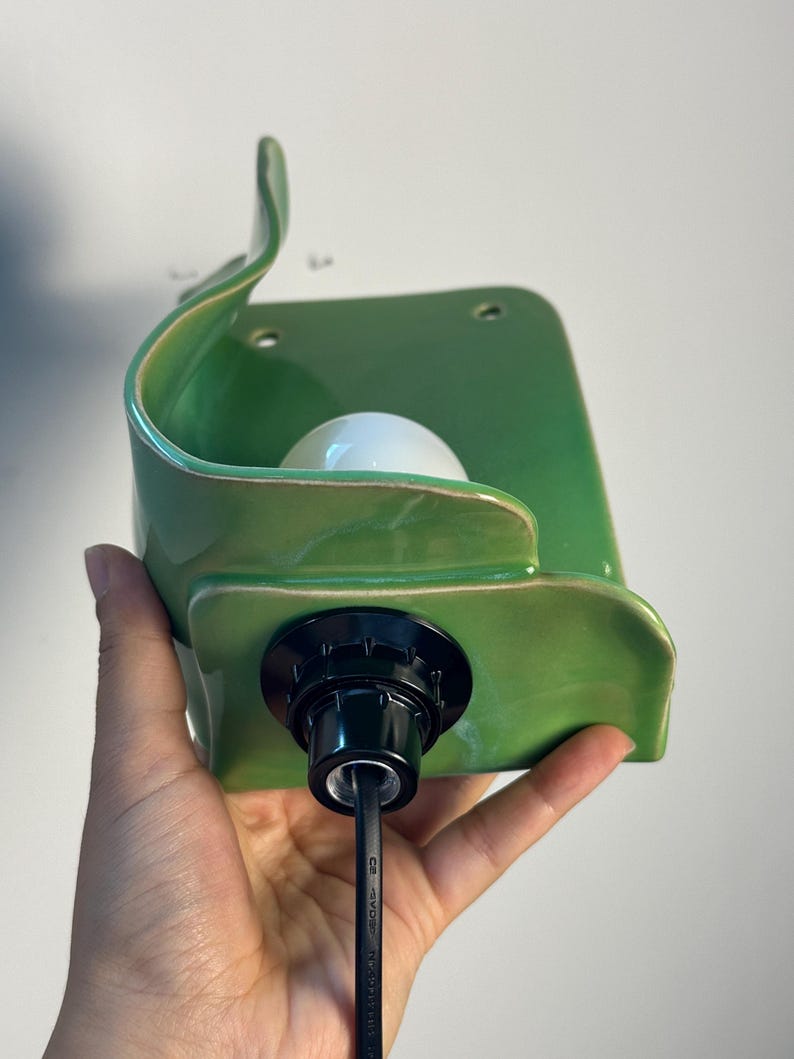 Pu&ograve; includere: Una lampada da parete in ceramica verde con una silhouette curva a forma di foglia e una finitura lucida. L'apparecchio presenta un portalampada centrale bianco e un cavo elettrico nero che esce dalla base, con due piccoli fori di montaggio.