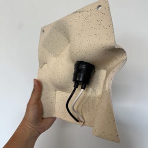 May include: A square, off-white wall lamp fixture with a speckled pattern. The surface has molded, undulating contours and two circular mounting holes at the top. A black plastic light socket is centered with one black and one white electrical wire.