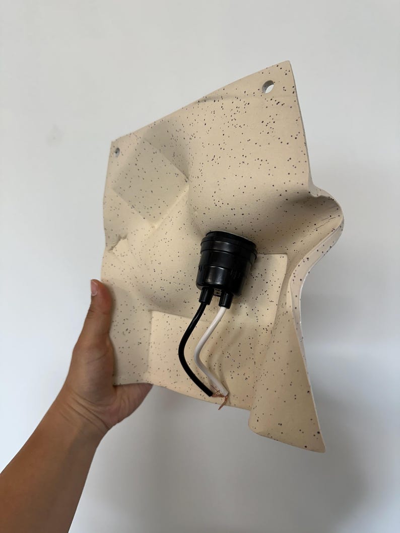 May include: A square, off-white wall lamp fixture with a speckled pattern. The surface has molded, undulating contours and two circular mounting holes at the top. A black plastic light socket is centered with one black and one white electrical wire.