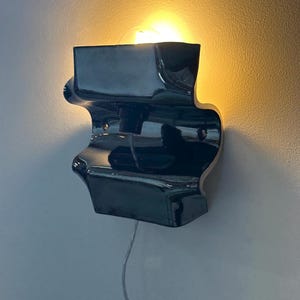May include: A glossy black ceramic wall sconce with a curved, undulating silhouette. The fixture features a central socket holding a glowing light bulb that casts a warm, soft light against the wall. A thin, clear power cord hangs from the bottom.