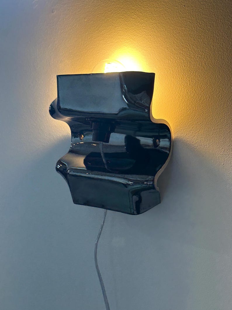 May include: A glossy black ceramic wall sconce with a curved, undulating silhouette. The fixture features a central socket holding a glowing light bulb that casts a warm, soft light against the wall. A thin, clear power cord hangs from the bottom.
