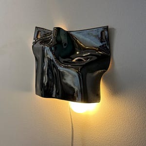 May include: A wall-mounted lamp featuring a glossy, dark charcoal-colored ceramic shade with an irregular, fluid, and undulating surface. The fixture emits a warm, glowing light from beneath the curved form, which is suspended by a thin cord.