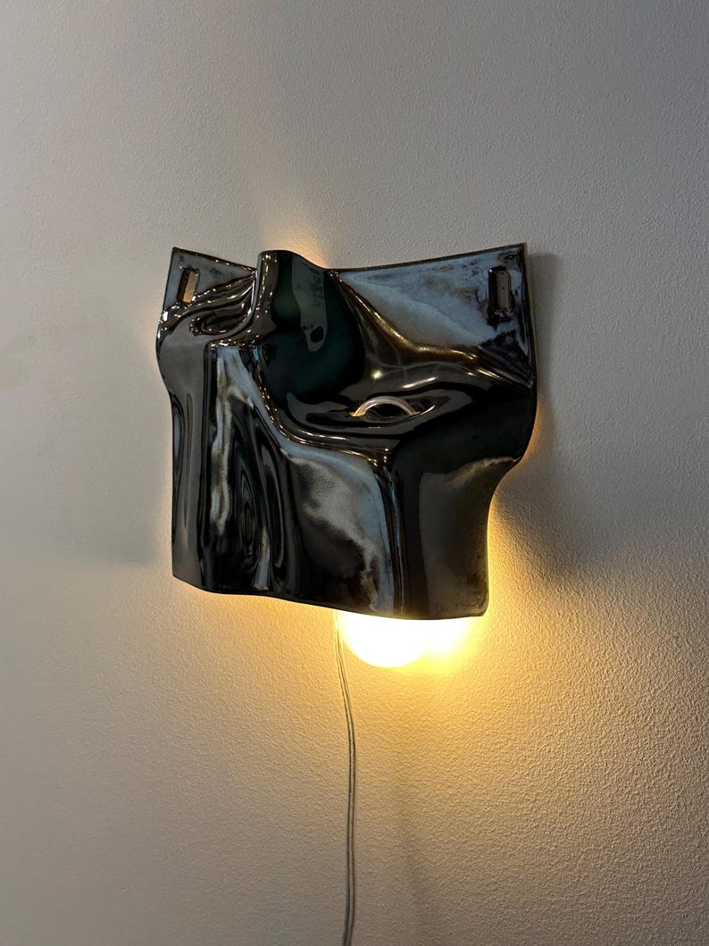 May include: A wall-mounted lamp featuring a glossy, dark charcoal-colored ceramic shade with an irregular, fluid, and undulating surface. The fixture emits a warm, glowing light from beneath the curved form, which is suspended by a thin cord.