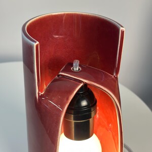 May include: A cylindrical ceramic table lamp with a glossy deep red glaze. The lamp features a unique cut-out design that exposes a black light socket and a white light bulb. A clear power cord is visible at the top of the structure.