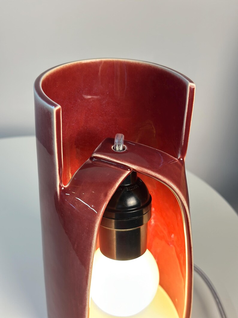 May include: A cylindrical ceramic table lamp with a glossy deep red glaze. The lamp features a unique cut-out design that exposes a black light socket and a white light bulb. A clear power cord is visible at the top of the structure.