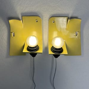 May include: A pair of wall-mounted sconces featuring curved, glossy yellow ceramic backplates with irregular edges. Each fixture includes a central black socket holding a round, frosted white light bulb, with a thin gray cord extending downward.