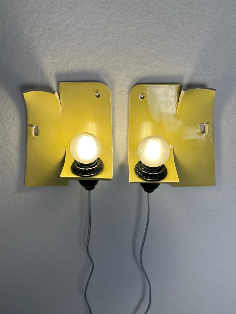 May include: A pair of wall-mounted sconces featuring curved, glossy yellow ceramic backplates with irregular edges. Each fixture includes a central black socket holding a round, frosted white light bulb, with a thin gray cord extending downward.