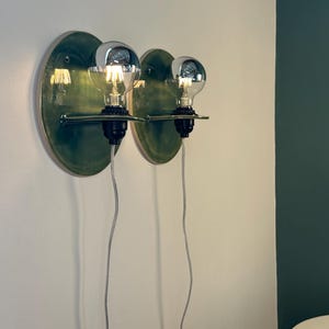 May include: Two wall-mounted light fixtures featuring circular, glossy green ceramic backplates. Each fixture has a black socket holding a clear, round light bulb with a silver-tinted top. A thin, horizontal metal arm supports each bulb, and a white cord hangs down from each base.