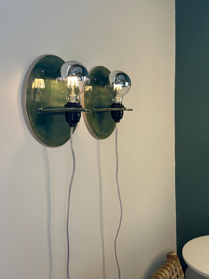 May include: Two wall-mounted light fixtures featuring circular, glossy green ceramic backplates. Each fixture has a black socket holding a clear, round light bulb with a silver-tinted top. A thin, horizontal metal arm supports each bulb, and a white cord hangs down from each base.
