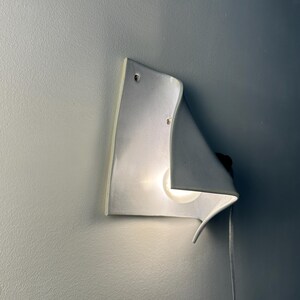 May include: A white ceramic wall sconce with a curved, folded sheet design. The fixture features a single glowing light bulb tucked behind the ceramic fold, casting a soft light. A thin white power cord extends from the side of the light fixture.