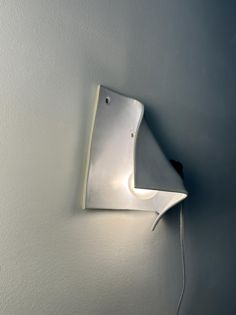 May include: A white ceramic wall sconce with a curved, folded sheet design. The fixture features a single glowing light bulb tucked behind the ceramic fold, casting a soft light. A thin white power cord extends from the side of the light fixture.