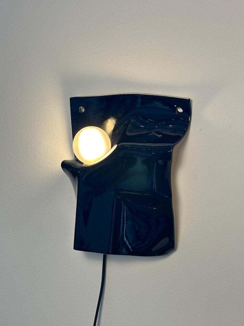 Puede incluir: Un aplique de pared con una base de cer&aacute;mica azul marino oscuro brillante de forma irregular y plegada. Una bombilla blanca redonda y brillante est&aacute; colocada dentro de una hendidura curva en el lado izquierdo de la superficie oscura y reflectante.