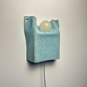 May include: A wall-mounted light fixture featuring a light blue ceramic base with a speckled finish. The base has a curved, rectangular shape with two mounting holes at the top corners and holds a single, round, frosted white glass light bulb.