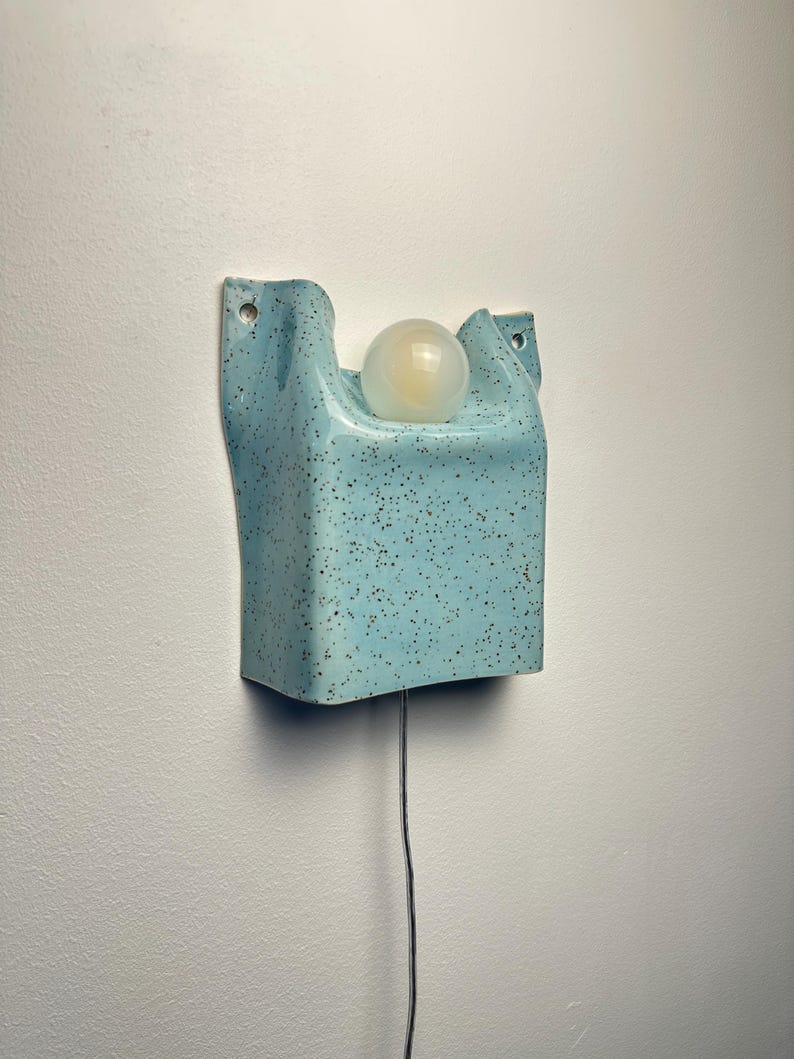 May include: A wall-mounted light fixture featuring a light blue ceramic base with a speckled finish. The base has a curved, rectangular shape with two mounting holes at the top corners and holds a single, round, frosted white glass light bulb.
