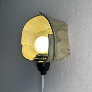 Puede incluir: Un aplique de pared con una pantalla de cer&aacute;mica curva de color amarillo p&aacute;lido y acabado brillante. Una bombilla blanca redonda y brillante est&aacute; centrada en la pantalla, unida a una base de casquillo negro con un cable transparente hacia abajo.