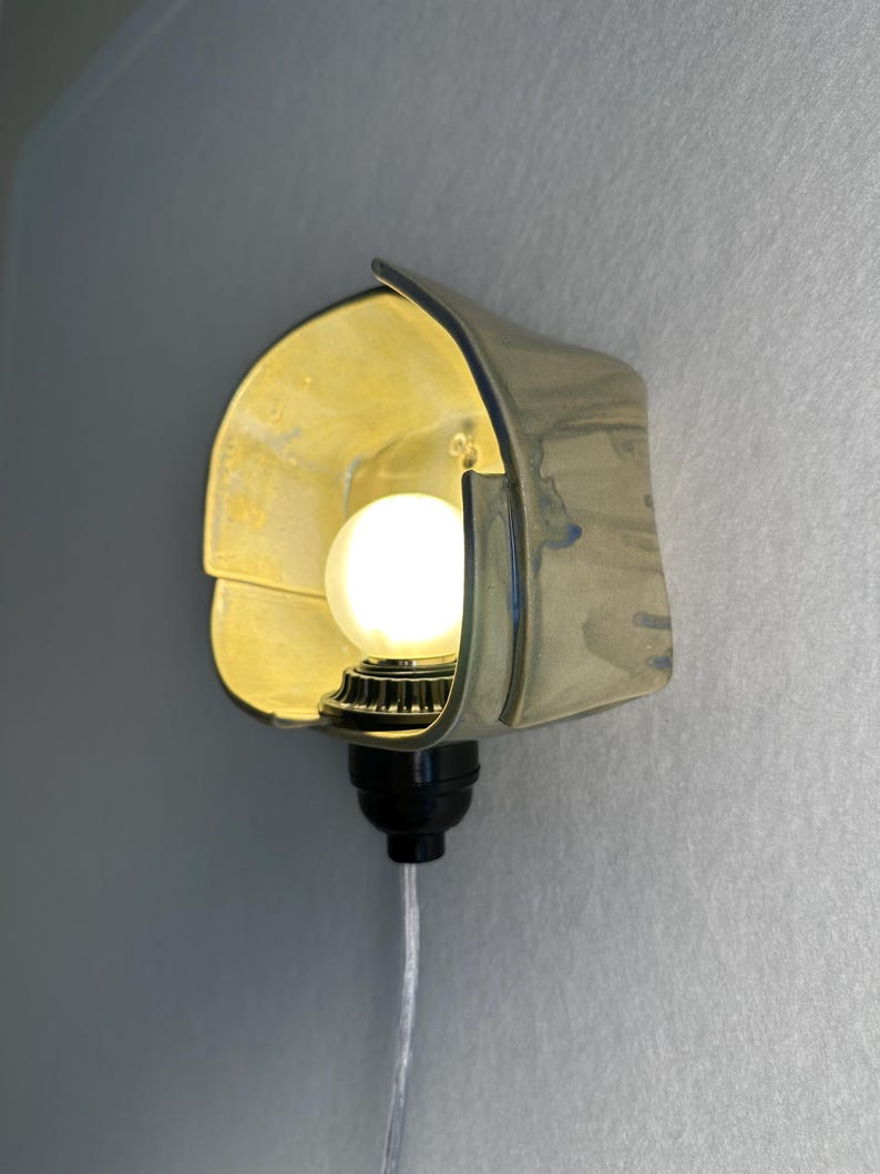 Puede incluir: Un aplique de pared con una pantalla de cer&aacute;mica curva de color amarillo p&aacute;lido y acabado brillante. Una bombilla blanca redonda y brillante est&aacute; centrada en la pantalla, unida a una base de casquillo negro con un cable transparente hacia abajo.