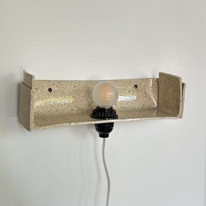 May include: A speckled beige ceramic wall sconce with a built-in shelf. The sconce has a black light fixture and a frosted bulb. The cord is gray. The sconce is mounted on a white wall.