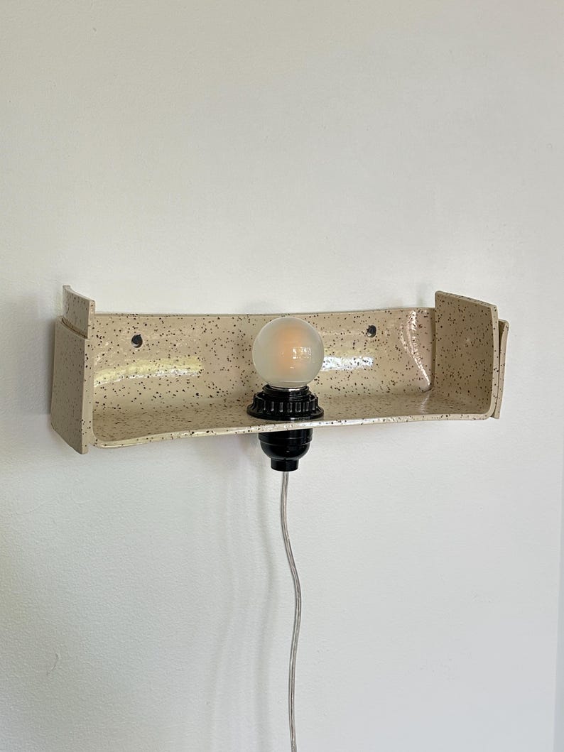May include: A speckled beige ceramic wall sconce with a built-in shelf. The sconce has a black light fixture and a frosted bulb. The cord is gray. The sconce is mounted on a white wall.