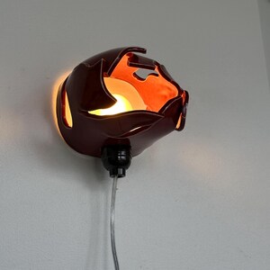 May include: A maroon wall sconce with an unusual, organic form. The sconce has a glossy finish and emits a warm, orange glow. It is connected to a black socket and a clear cord. The sconce is mounted on a white wall.