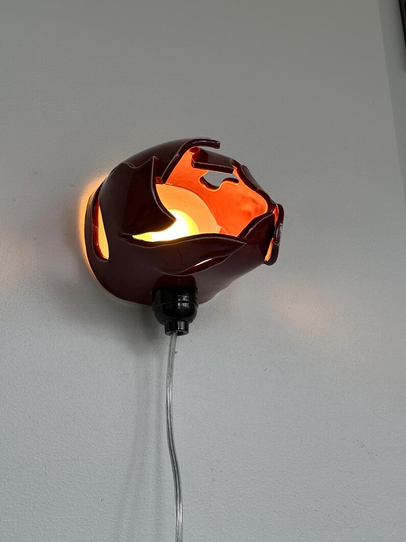 May include: A maroon wall sconce with an unusual, organic form. The sconce has a glossy finish and emits a warm, orange glow. It is connected to a black socket and a clear cord. The sconce is mounted on a white wall.