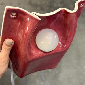Puede incluir: Un aplique de pared de cer&aacute;mica con una superficie roja oscura brillante y bordes ondulados. La l&aacute;mpara cuenta con una bombilla esf&eacute;rica de cristal blanco esmerilado en el centro y dos orificios pretaladrados en la parte superior para su montaje.