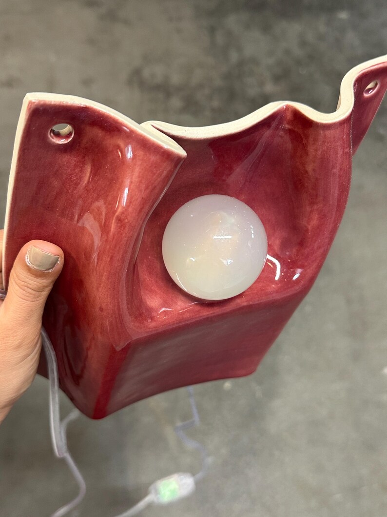 Puede incluir: Un aplique de pared de cer&aacute;mica con una superficie roja oscura brillante y bordes ondulados. La l&aacute;mpara cuenta con una bombilla esf&eacute;rica de cristal blanco esmerilado en el centro y dos orificios pretaladrados en la parte superior para su montaje.