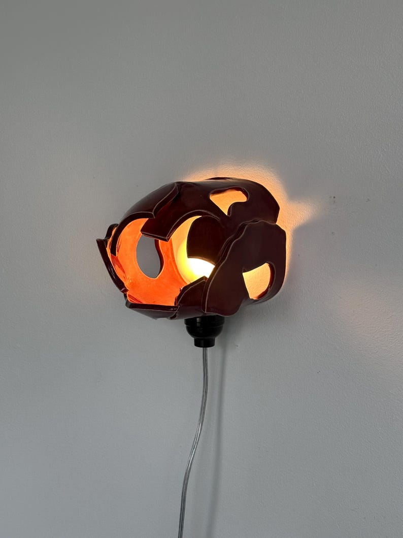 May include: A distinctive, dark brown wall sconce with an organic, cut-out design. The interior glows with warm orange light, creating a striking contrast. The sconce is mounted on a grey wall and has a black base with a silver cord.